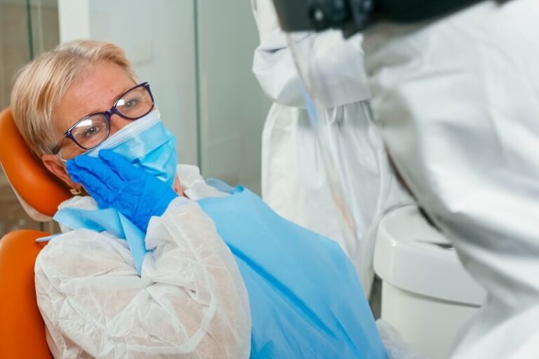 patient-with-protection-suit-touching-face-having-toothache-stomatology-clinic-global-pandemic-old-woman-explaining-dental-problem-doctor-dressed-coverall-with-face-shield-glovel (1) استبدال زراعة الأسنان من NHS